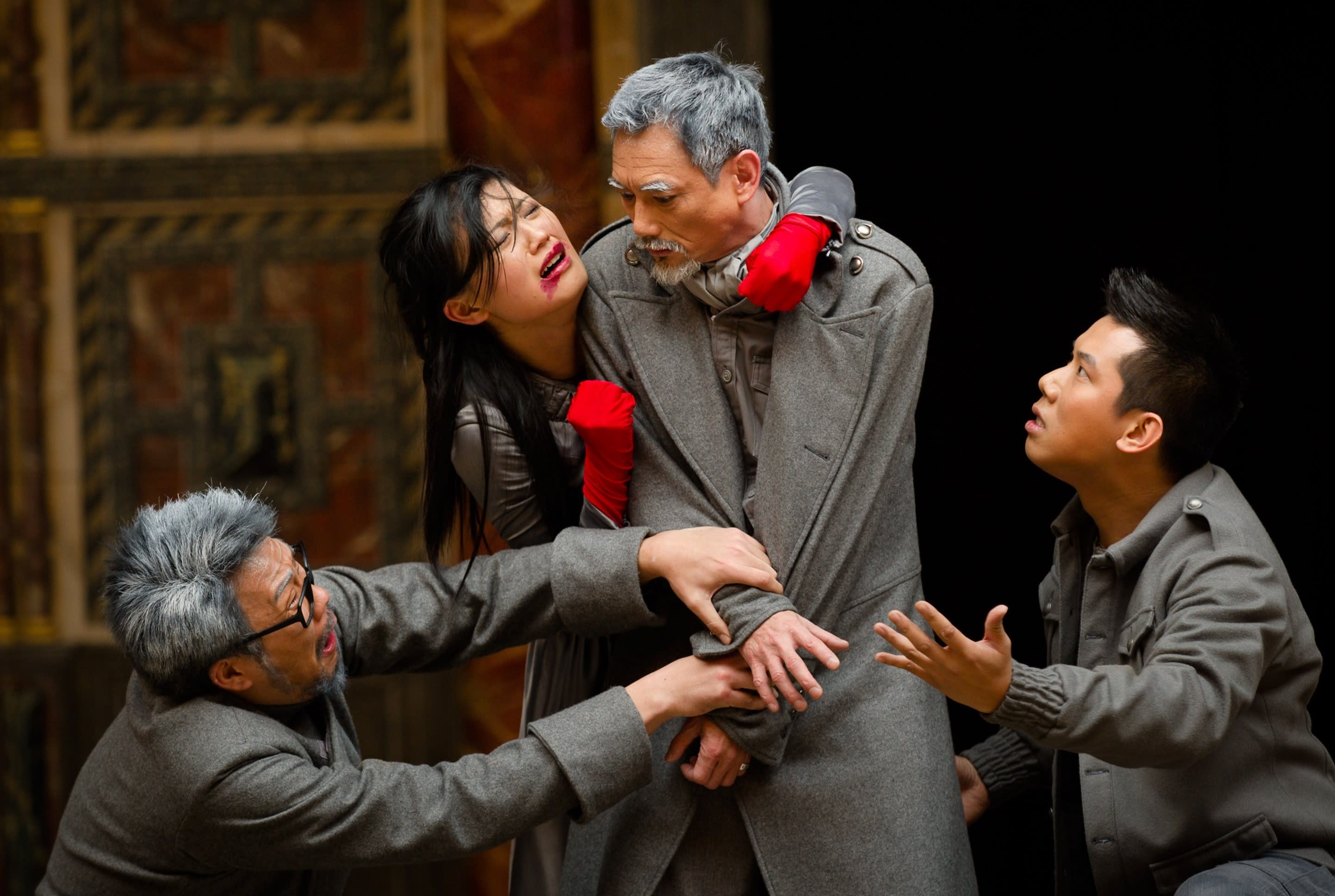Two men wearing grey coats look up at a man in a grey coat who has a grey beard, one man holds his wrists, and a woman grasps him from behind with an anguished expression